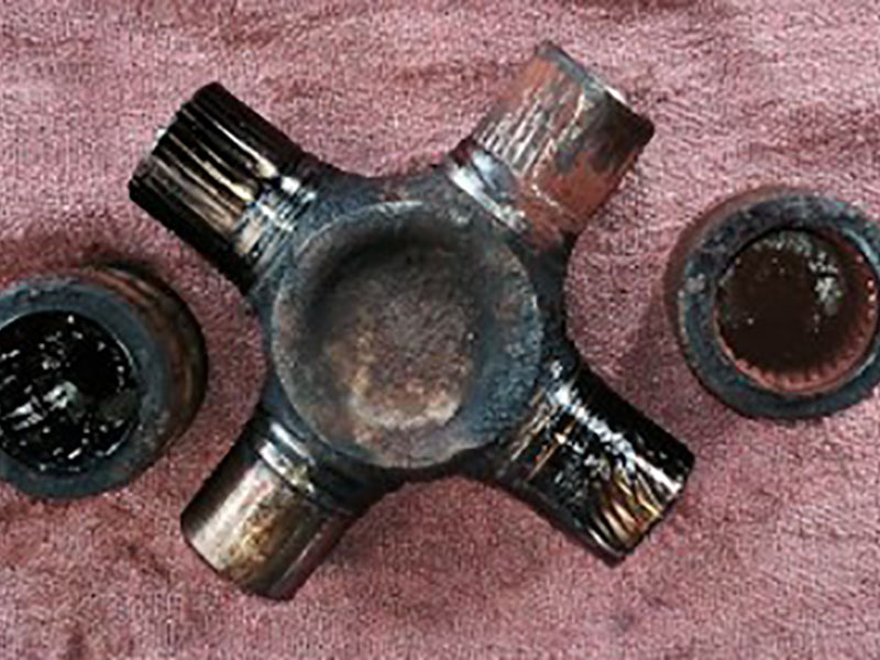 Worn universal joint cross and bearing cups showing common failure causes like lack of lubrication and excessive operating angles on a driveshaft